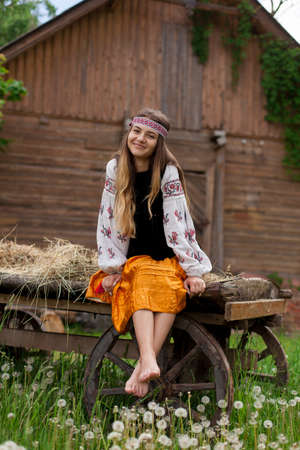 Beautiful slender Ukrainian woman in embroidery on the background of a wooden frame of a barn in the village sits on a wooden cartの写真素材