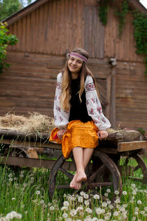 Beautiful slender Ukrainian woman in embroidery on the background of a wooden frame of a barn in the village sits on a wooden cartの写真素材