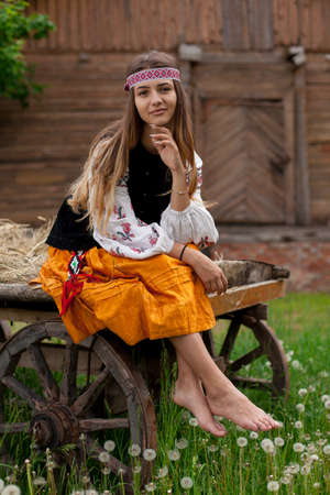 Beautiful slender Ukrainian woman in embroidery on the background of a wooden frame of a barn in the village sits on a wooden cartの写真素材