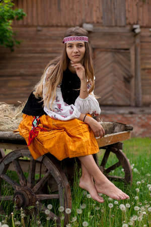 Beautiful slender Ukrainian woman in embroidery on the background of a wooden frame of a barn in the village sits on a wooden cartの写真素材