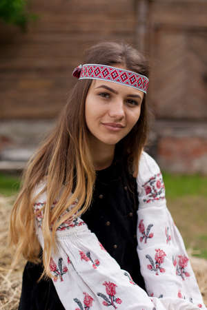 Beautiful slender Ukrainian woman in embroidery on the background of a wooden frame of a barn in the village sits on a wooden cartの写真素材