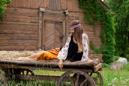 Beautiful slender Ukrainian woman in embroidery on the background of a wooden frame of a barn in the village sits on a wooden cartの写真素材
