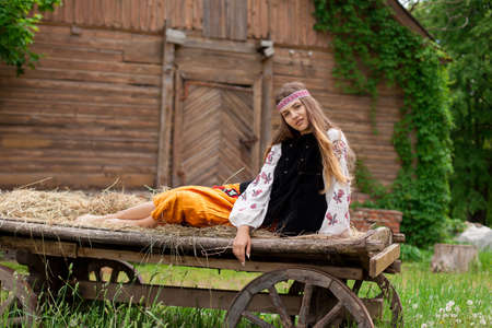 Beautiful slender Ukrainian woman in embroidery on the background of a wooden frame of a barn in the village sits on a wooden cartの写真素材