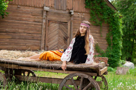 Beautiful slender Ukrainian woman in embroidery on the background of a wooden frame of a barn in the village sits on a wooden cartの写真素材