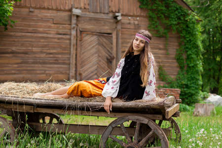 Beautiful slender Ukrainian woman in embroidery on the background of a wooden frame of a barn in the village sits on a wooden cartの写真素材