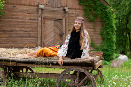 Beautiful slender Ukrainian woman in embroidery on the background of a wooden frame of a barn in the village sits on a wooden cartの写真素材