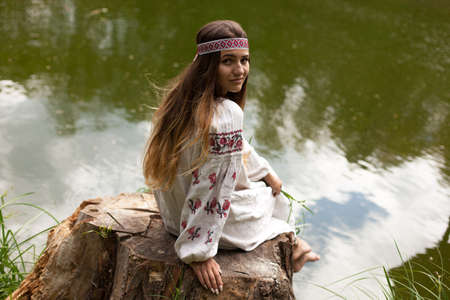 Beautiful slender Ukrainian in embroidery on the shore of the pond enjoys the nature of a summer sunny dayの写真素材