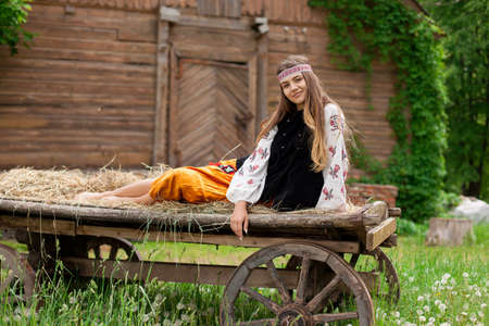Beautiful slender Ukrainian woman in embroidery on the background of a wooden frame of a barn in the village sits on a wooden cartの写真素材