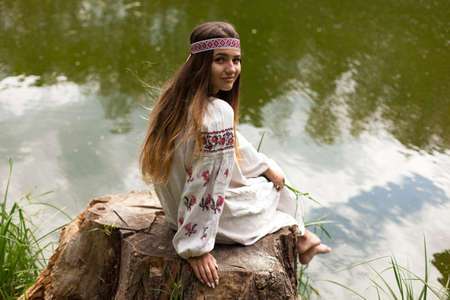 A beautiful Ukrainian woman dressed in embroidery sits on a stump on the shore of a lakeの写真素材