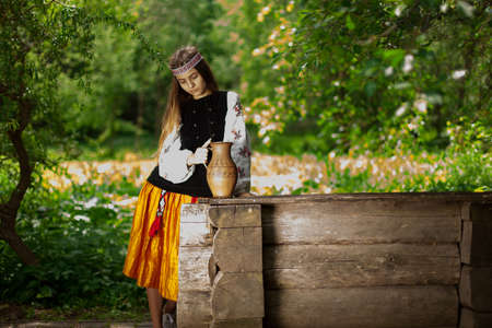Beautiful slender Ukrainian in embroidery on the background of a wooden well gaining fresh waterの写真素材