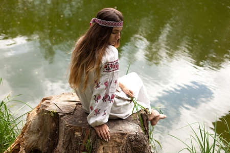 A beautiful Ukrainian woman dressed in embroidery sits on a stump on the shore of a lakeの写真素材