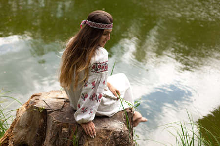 A beautiful Ukrainian woman dressed in embroidery sits on a stump on the shore of a lakeの写真素材