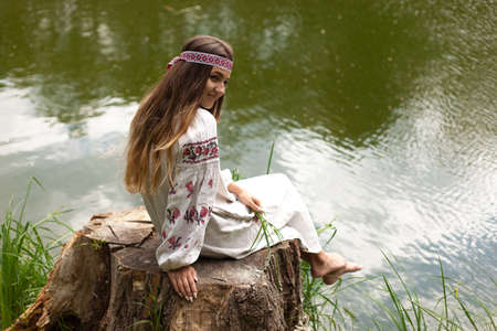 Beautiful slender Ukrainian in embroidery on the shore of the pond enjoys the nature of a summer sunny dayの写真素材
