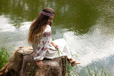 Beautiful slender Ukrainian in embroidery on the shore of the pond enjoys the nature of a summer sunny dayの写真素材