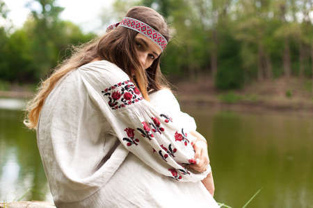 A beautiful Ukrainian woman dressed in embroidery sits on a stump on the shore of a lakeの写真素材