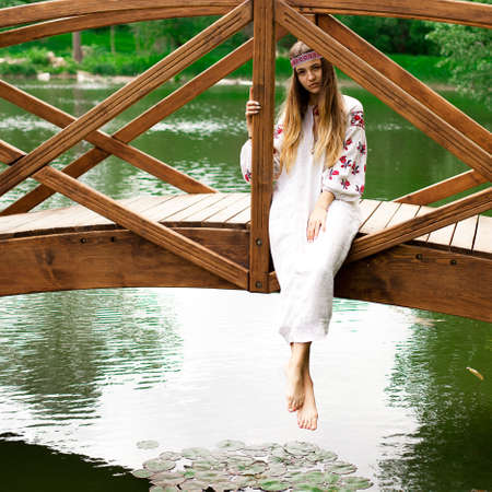 Beautiful slender Ukrainian in embroidery sits on the bridge over the waterの写真素材