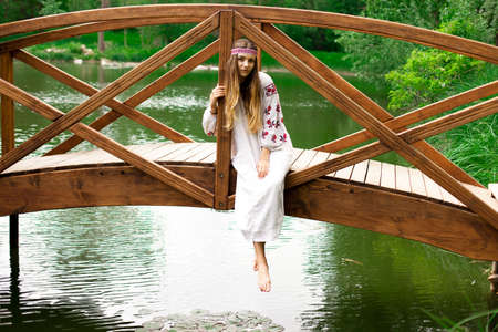Beautiful Ukrainian woman wearing embroidery sits on a wooden bridge on a pondの写真素材