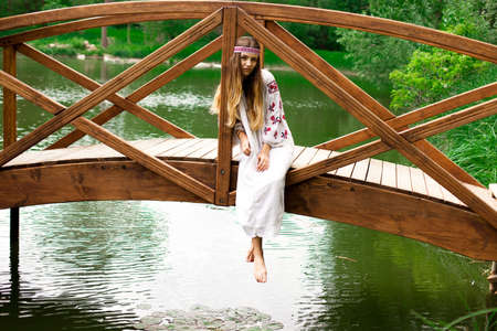 Beautiful Ukrainian woman wearing embroidery sits on a wooden bridge on a pondの写真素材