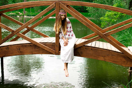 Beautiful slender Ukrainian in embroidery sits on the bridge over the waterの写真素材