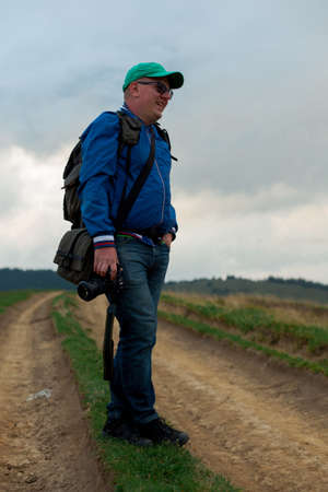 photographer in blue windbreaker and jeans with a camera and a backpack is standing on a dirt road in the mountainsの写真素材