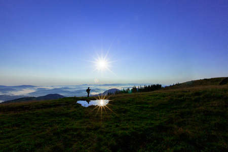 the top of the mountain with the silhouette of a man and the silhouettes of the pines opposite the bright blue sky and the blue peaks of the mountains in the distance, the sun's rays directly into the lens and reflected in a puddleの写真素材