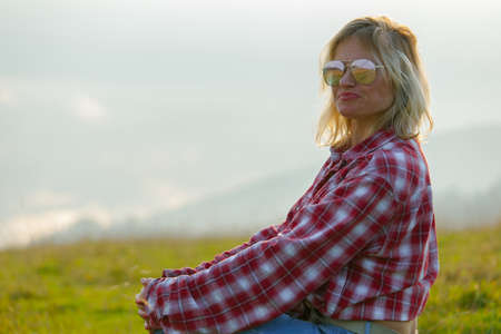 blonde woman in denim clothes and sunglasses sits on a meadow in the foggy mountainsの写真素材
