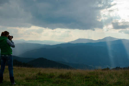 girl photographer in the mountains shoots the landscape on the background of the sun's rays on a cloudy dayの写真素材