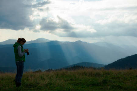 girl photographer in the mountains shoots the landscape on the background of the sun's rays on a cloudy dayの写真素材