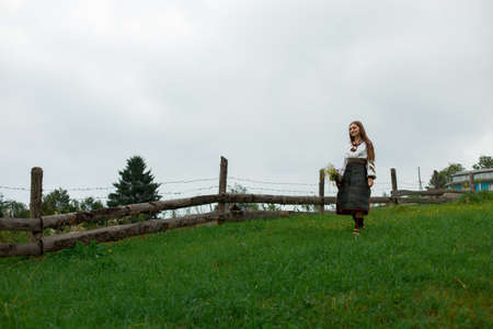 girl in embroidery with a bouquet of wildflowers walks along the green grass along a wooden fenceの写真素材