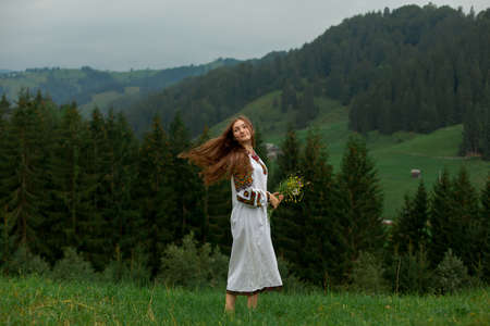 girl with loose hair in an embroidery with a bouquet of wildflowers happily runs along the green in the carpathian mountainsの写真素材