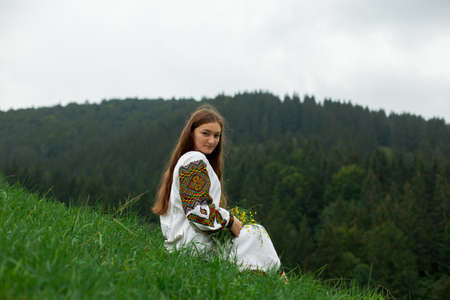girl with loose hair in an embroidery with a bouquet of wild flowers sits on green grass against the background of the carpathian mountainsの写真素材