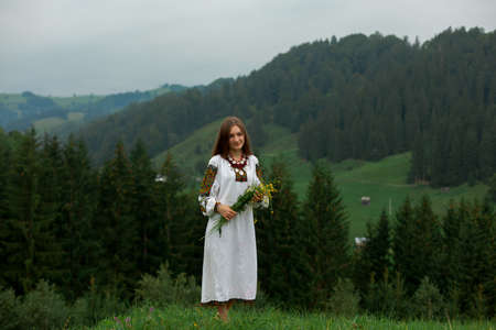 girl in embroidery with a bouquet of wild flowers stands on green grass in the carpathian mountainsの写真素材