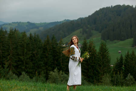 girl with loose hair in an embroidery with a bouquet of wildflowers happily runs along the green in the carpathian mountainsの写真素材
