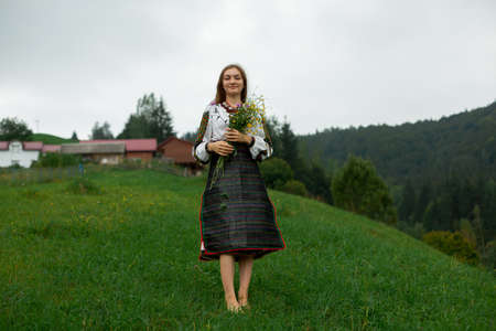girl in embroidery with a bouquet of wild flowers stands on green grass in the carpathian mountainsの写真素材