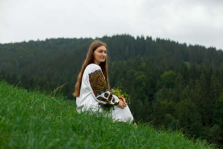 girl with loose hair in an embroidery with a bouquet of wild flowers sits on green grass against the background of the carpathian mountainsの写真素材