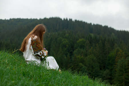 girl in embroidery with a bouquet of wild flowers stands on green grass in the carpathian mountainsの写真素材