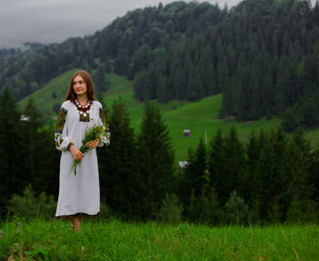 girl in embroidery with a bouquet of wild flowers stands barefoot on green grass in the carpathian mountainsの写真素材
