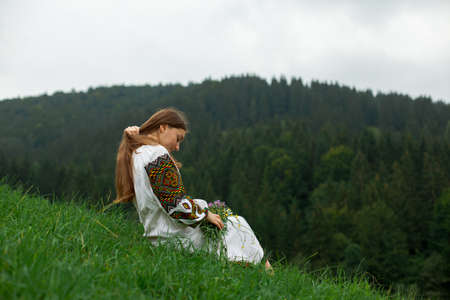 girl in embroidery with a bouquet of wild flowers stands on green grass in the carpathian mountainsの写真素材
