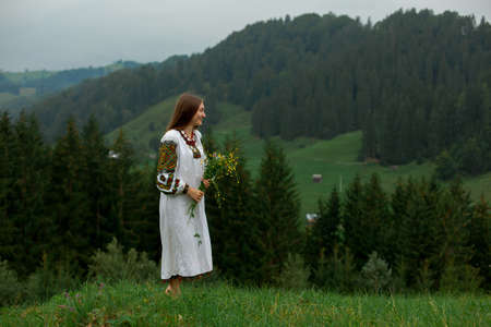 girl in embroidery with a bouquet of wild flowers stands on green grass in the carpathian mountainsの写真素材