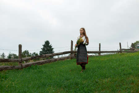 girl in embroidery with a bouquet of wild flowers stands on green grass in the carpathian mountainsの写真素材