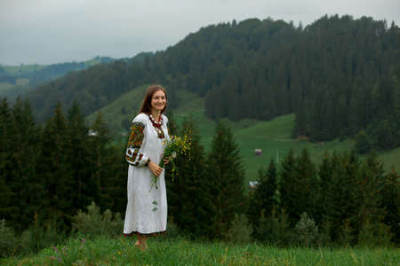 girl in embroidery with a bouquet of wild flowers stands on green grass in the carpathian mountainsの写真素材