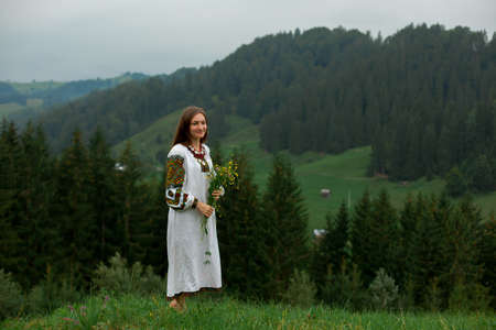 girl in embroidery with a bouquet of wild flowers stands on green grass in the carpathian mountainsの写真素材