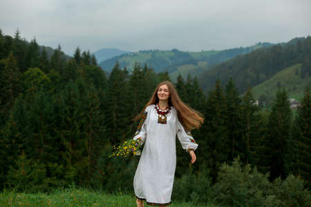 girl in embroidery with a bouquet of wild flowers stands on green grass in the carpathian mountainsの写真素材