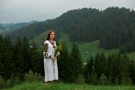 girl in embroidery with a bouquet of wild flowers stands on green grass in the carpathian mountainsの写真素材