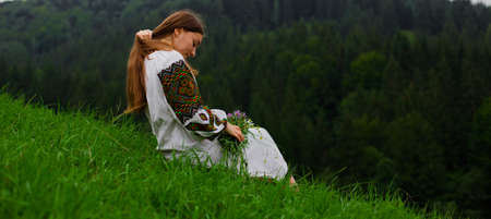 girl in embroidery with a bouquet of wild flowers stands on green grass in the carpathian mountainsの写真素材