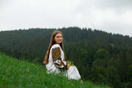 girl in embroidery with a bouquet of wild flowers stands on green grass in the carpathian mountainsの写真素材