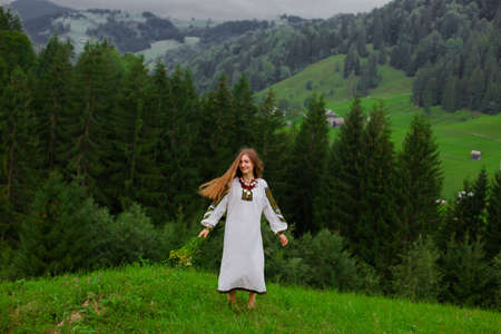 girl in embroidery with a bouquet of wild flowers stands on green grass in the carpathian mountainsの写真素材