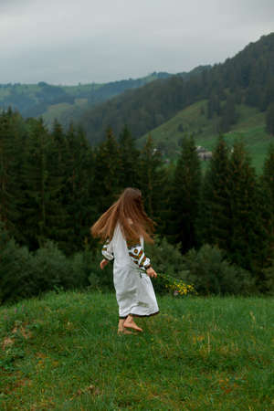 girl in embroidery with a bouquet of wild flowers stands on green grass in the carpathian mountainsの写真素材