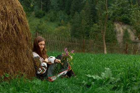 girl in embroidery with a bouquet of wild flowers stands on green grass in the carpathian mountainsの写真素材