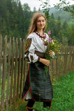 girl in embroidery with a bouquet of wild flowers stands on green grass in the carpathian mountainsの写真素材